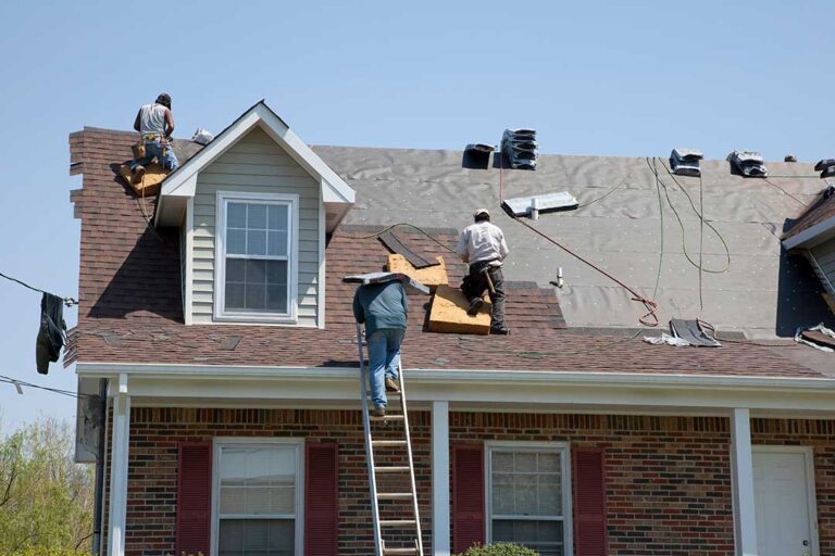 Roofing material samples arranged beside a home with a steep roof after light snowfall.